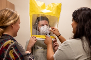 Woman wears a yellow tent-like bag with clear screen that includes a breathing apparatus while another person assists her with the equipment 