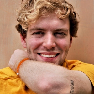 Young man with wavy blonde hair wears a yellow shirt and shows a tattoo on his arm that says "BRAVE"