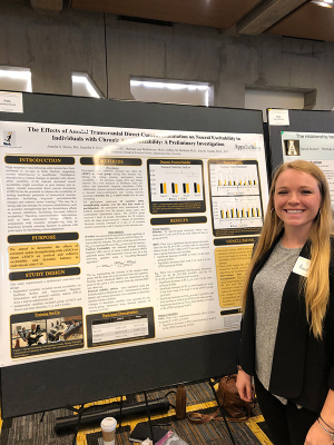 Young woman with long, blonde hair stands beside a research poster with different text boxes and writing on it 