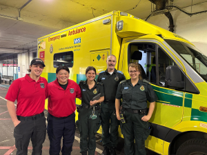 Group of five people wearing official looking logoed shirts stand side by side in front of a yellow ambulance 