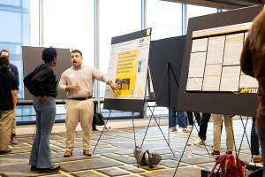 Person points to a poster on an easel while talking to someone else in a large room with other posters. 