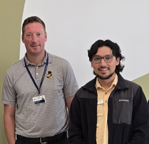 Two people stand in front of a green wall with a white graphic; one is wearing a lanyard and a name badge