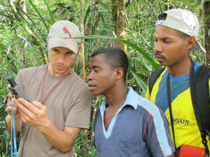 Man holds an instrument in a forested area with two other men looking over his shoulder 