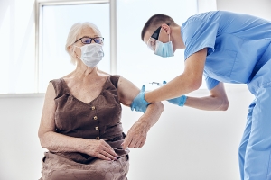Person in scrubs leans over to give a shot to an older woman sitting in a chair 