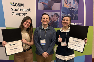 Two women hold certificates with a man standing between them and a banner behind them that reads SEACSM conference